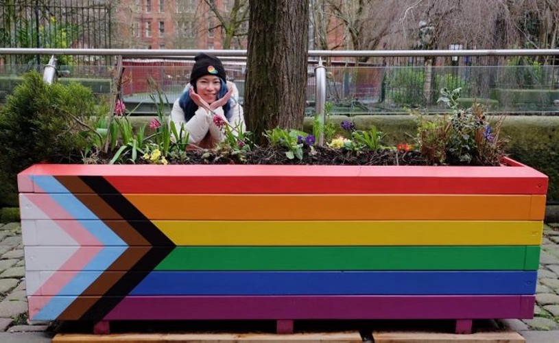 LGBTQ+ activist Qiuyan Chen posing behind a rainbow flag flower bed